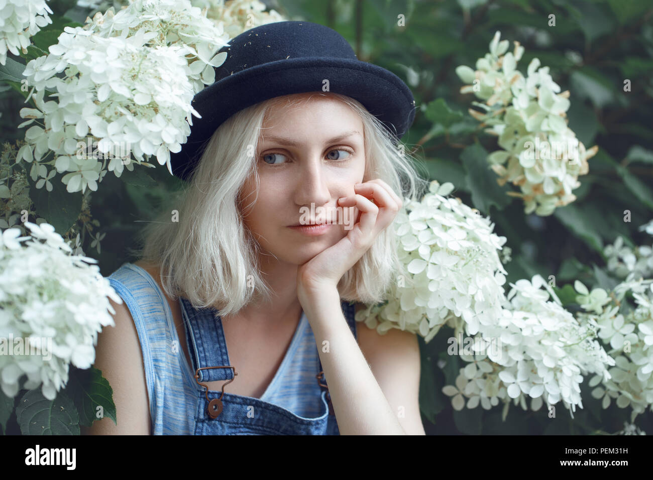 Closeup portrait of beautiful Caucasian teenage young blonde model girl woman in blue tshirt, jeans romper, black hat, sitting among large white flowe Stock Photo