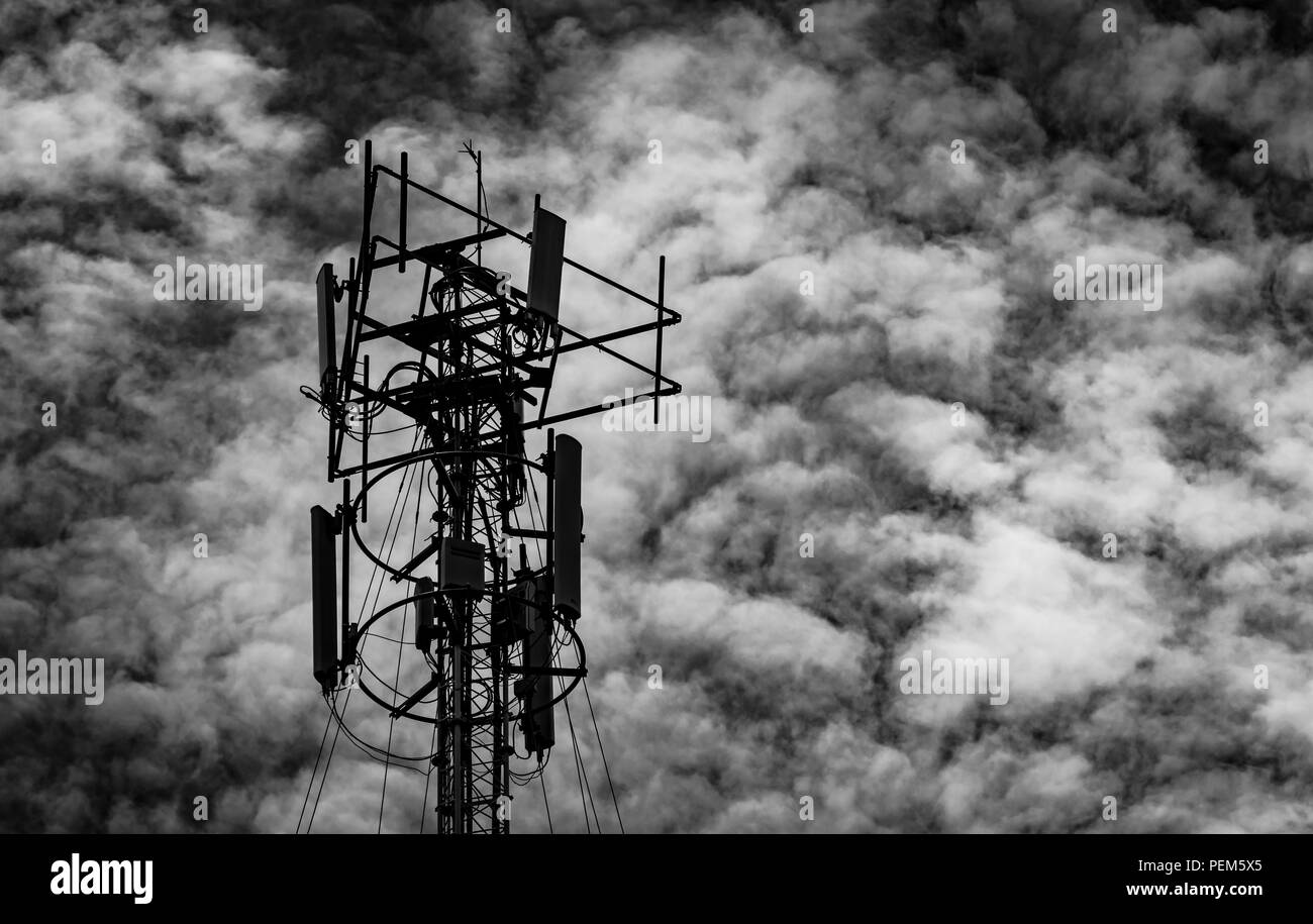 Black and white picture of telecommunication tower against grey sky and white clouds. Antenna on dark sky background. Radio and satellite pole. Commun Stock Photo