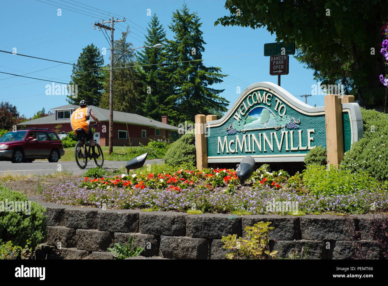 A roadside sign welcoming people to the city of McMinnville surrounded by planted flowers. A car and a bicyclist are passing by Stock Photo