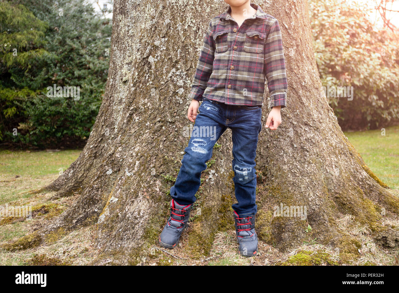 stylish little boy in casual clothes jean and shirt is posing with big tree in background PER32H
