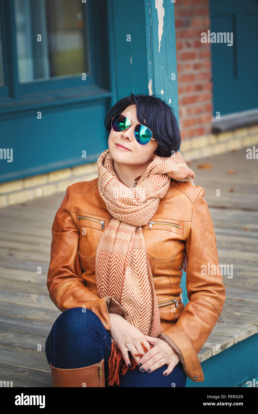 Portrait of smiling middle aged caucasian dark haired brunette woman with short bob hair  in sunglasses wearing light brown leather jacket and scarf o Stock Photo