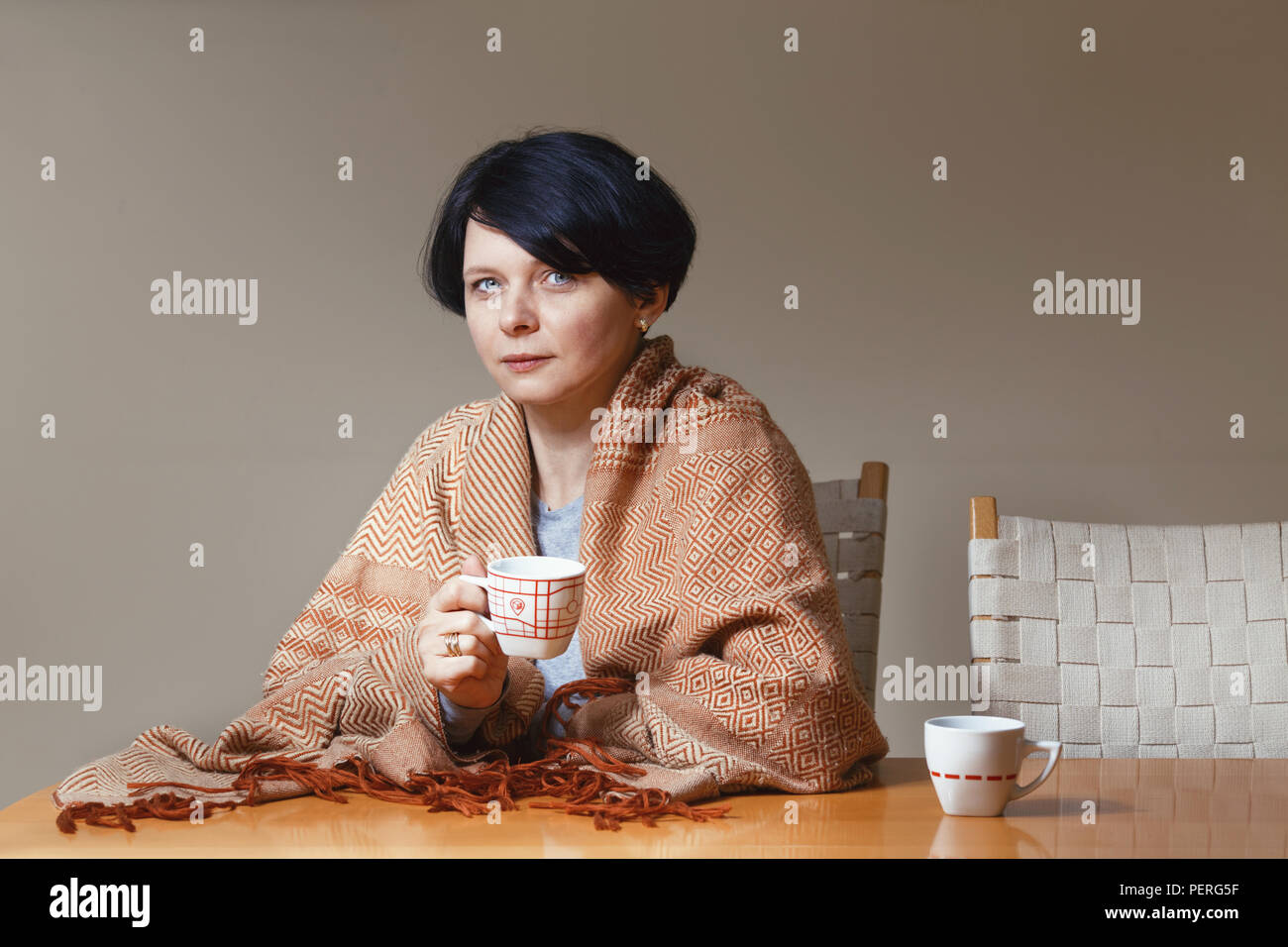 Portrait of beautiful Caucasian brunette mid age woman covered with blanket sitting at table drinking tea coffee holding cup in hands looking directly Stock Photo