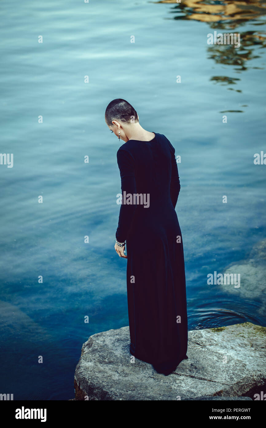 Portrait of sad beautiful Caucasian white young bald girl woman with shaved hair head standing by water lake looking away, mystic dark mood, toned wit Stock Photo