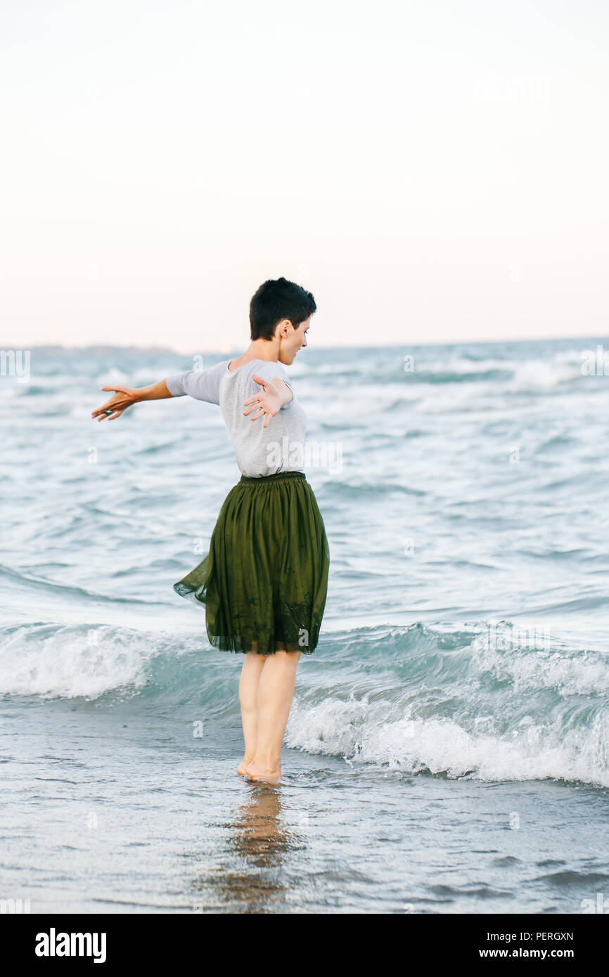 Portrait of beautiful Caucasian white brunette woman with short hair in grey shirt, green olive tutu tulle skirt, standing barefoot on beach in sea wa Stock Photo