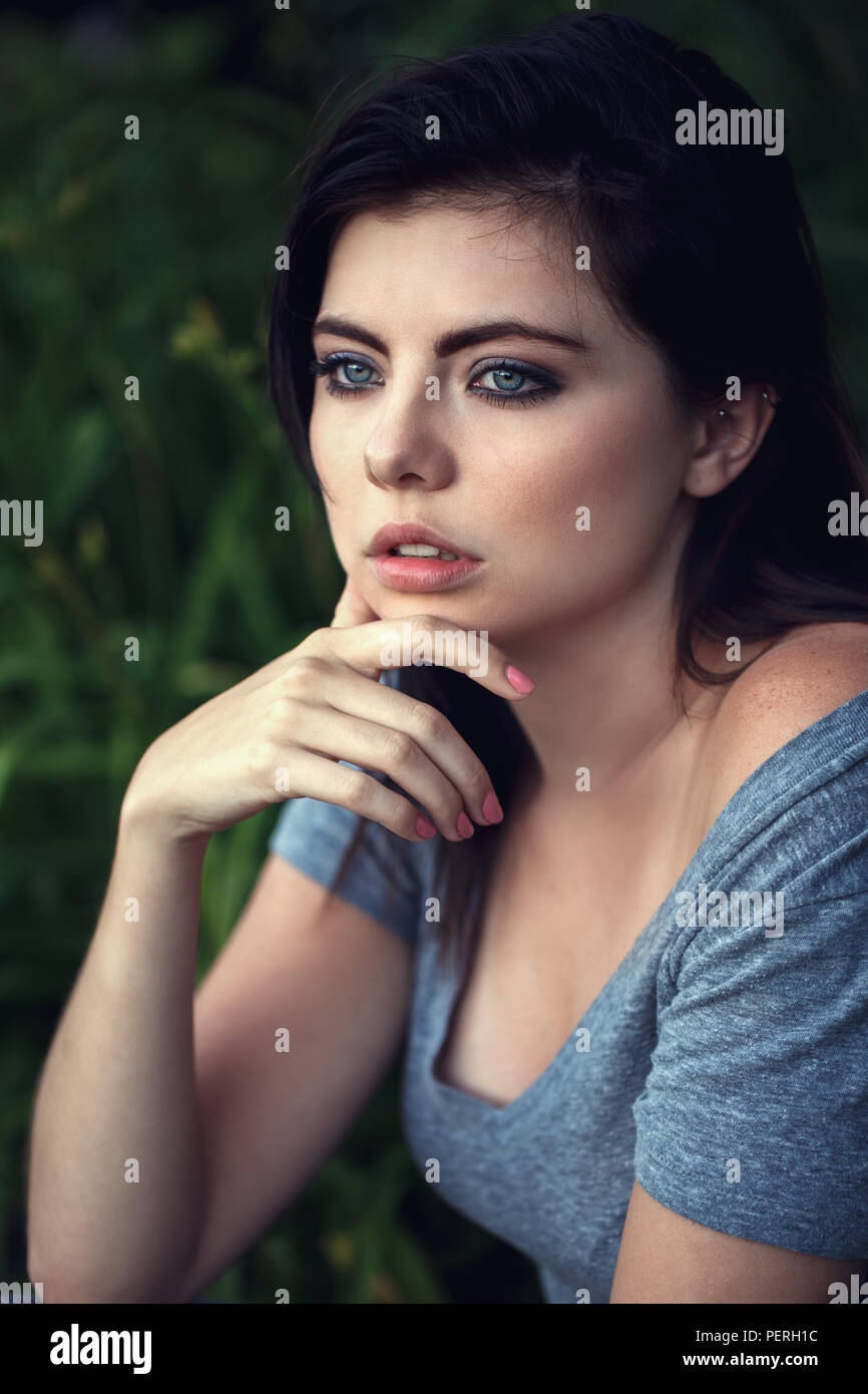 Closeup portrait of beautiful sexy young Caucasian woman with black hair, blue eyes, looking away, sitting outdoors, in grey open tshirt, natural beau Stock Photo