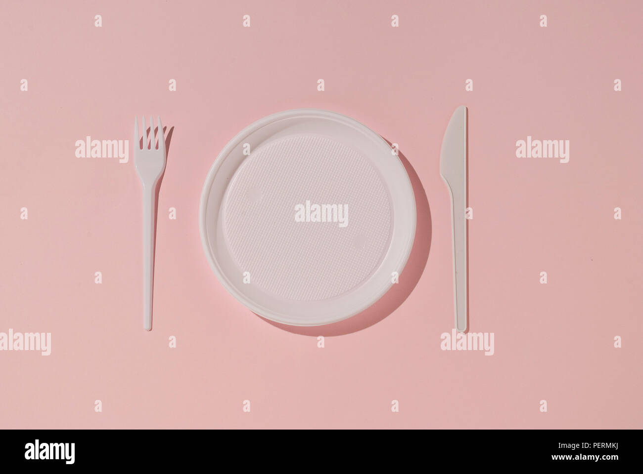 White plastic plate, knife and fork on a pink background. Plastic tableware. Stock Photo
