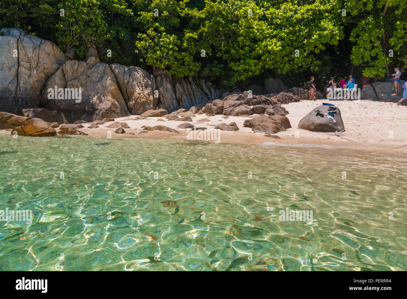 After a snorkelling trip, tourists are eating at the rocky part of Rawa beach under the shade of trees, while the fishes are swimming in the shallow... Stock Photo