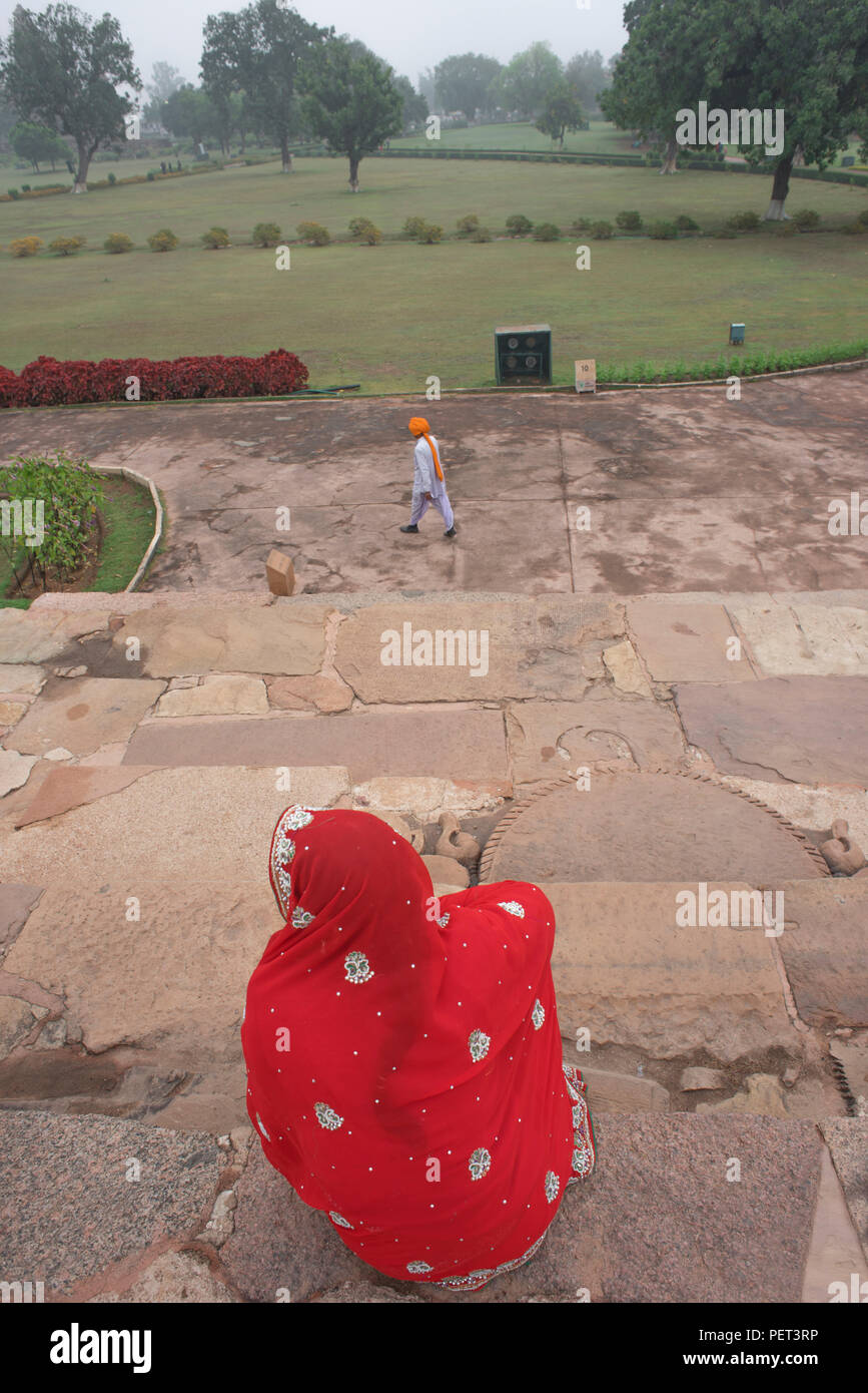 An Indian lady sitting on the steps of a temple wearing a red saree  and a man in an orange headgear and white dress walking away from her in a park Stock Photo
