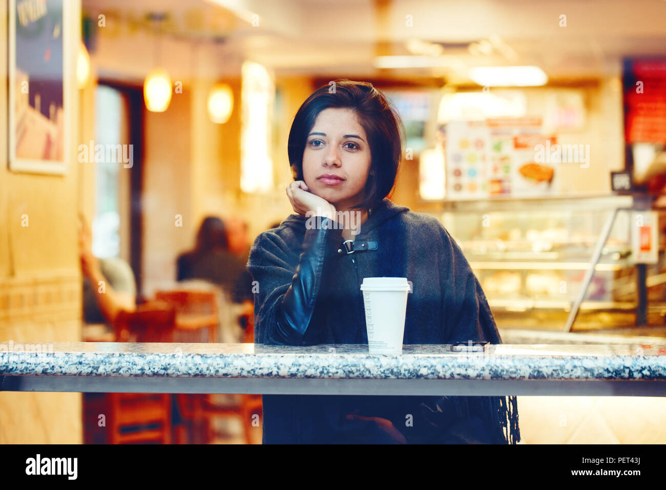 Candid portrait of beautiful young hipster latin hispanic girl woman with short hair bob, in grey cape poncho jacket, sitting in cafe restaurant with  Stock Photo