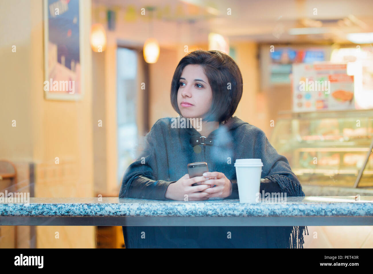 Candid portrait of beautiful young hipster latin hispanic girl woman with short hair bob in grey poncho jacket sitting in cafe  with coffee and cell p Stock Photo