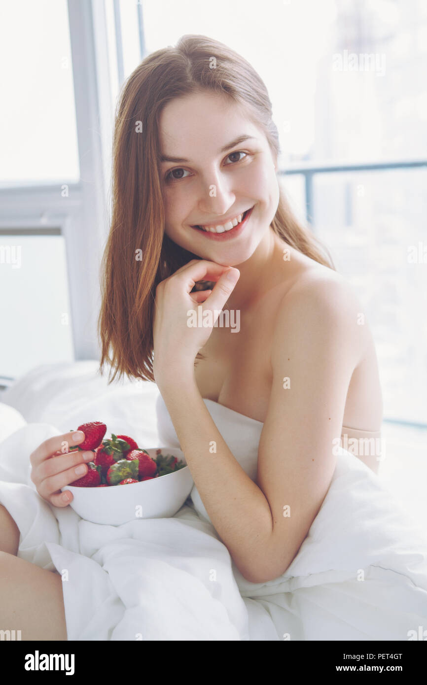 Portrait of smiling beautiful young Caucasian woman with long hair sitting in bed early morning by window, eating red fresh strawberry, lifestyle, ton Stock Photo