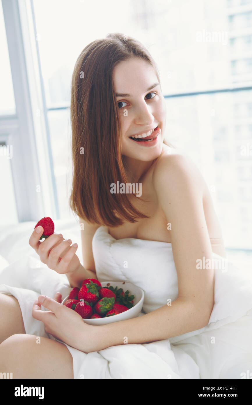 Portrait of smiling beautiful young Caucasian woman with long hair sitting in bed early morning by window, eating red fresh strawberry, showing her to Stock Photo