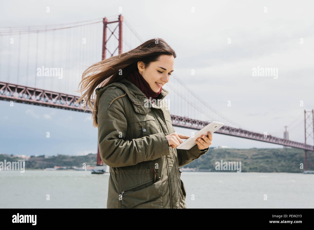 A young beautiful woman uses a tablet to communicate with friends or looks at a map or calls a taxi or something else. Bridge April 25 in Lisbon in Portugal in the background. Stock Photo