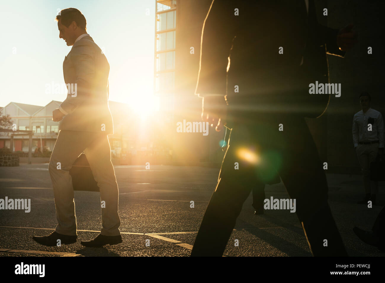 Businessmen in hurry to reach office walking on city street with sun flare in the background. Men commuting to office early in the morning carrying of Stock Photo
