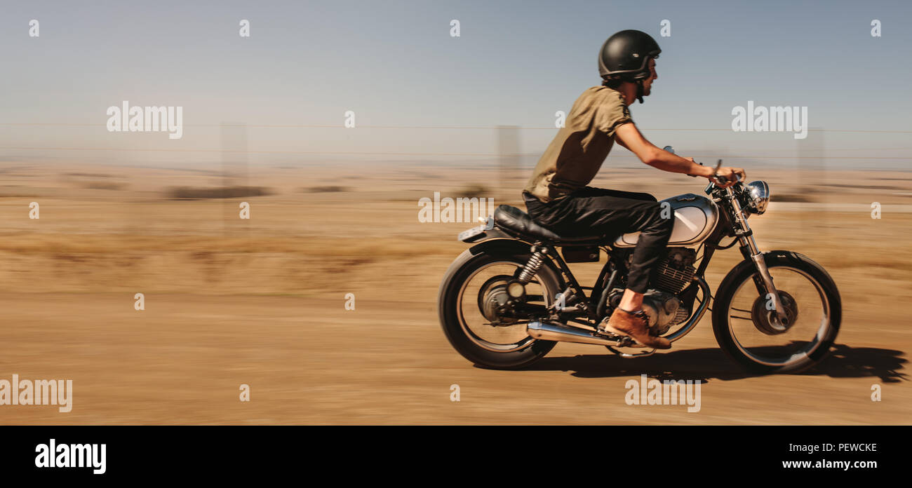Side view of young man biker riding his bike on country road. Male rider driving motorcycle on a dirt road in country side. Stock Photo