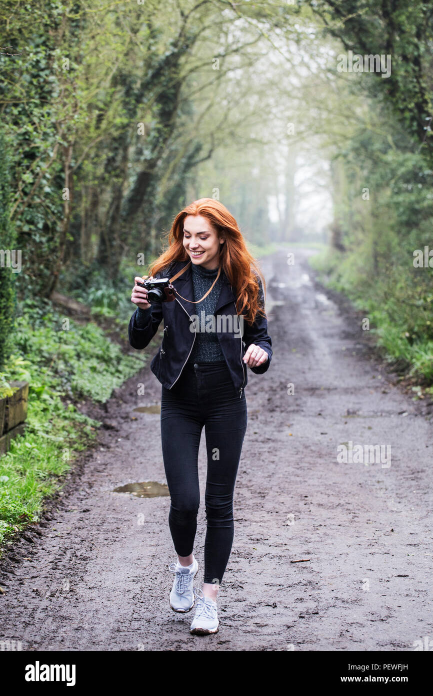 Smiling young woman with long red hair walking along forest path ...