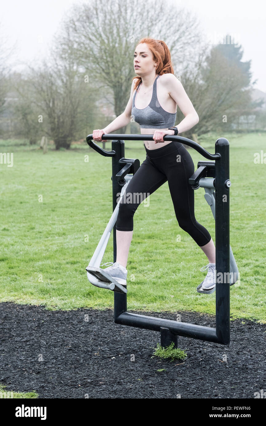 Young woman with long red hair wearing sportswear, using outdoor exercise machine. Stock Photo