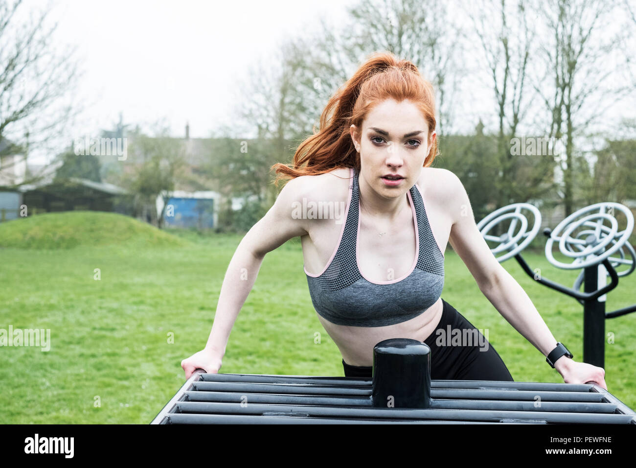 Young woman with long red hair wearing sportswear, using outdoor exercise machine, looking at camera. Stock Photo