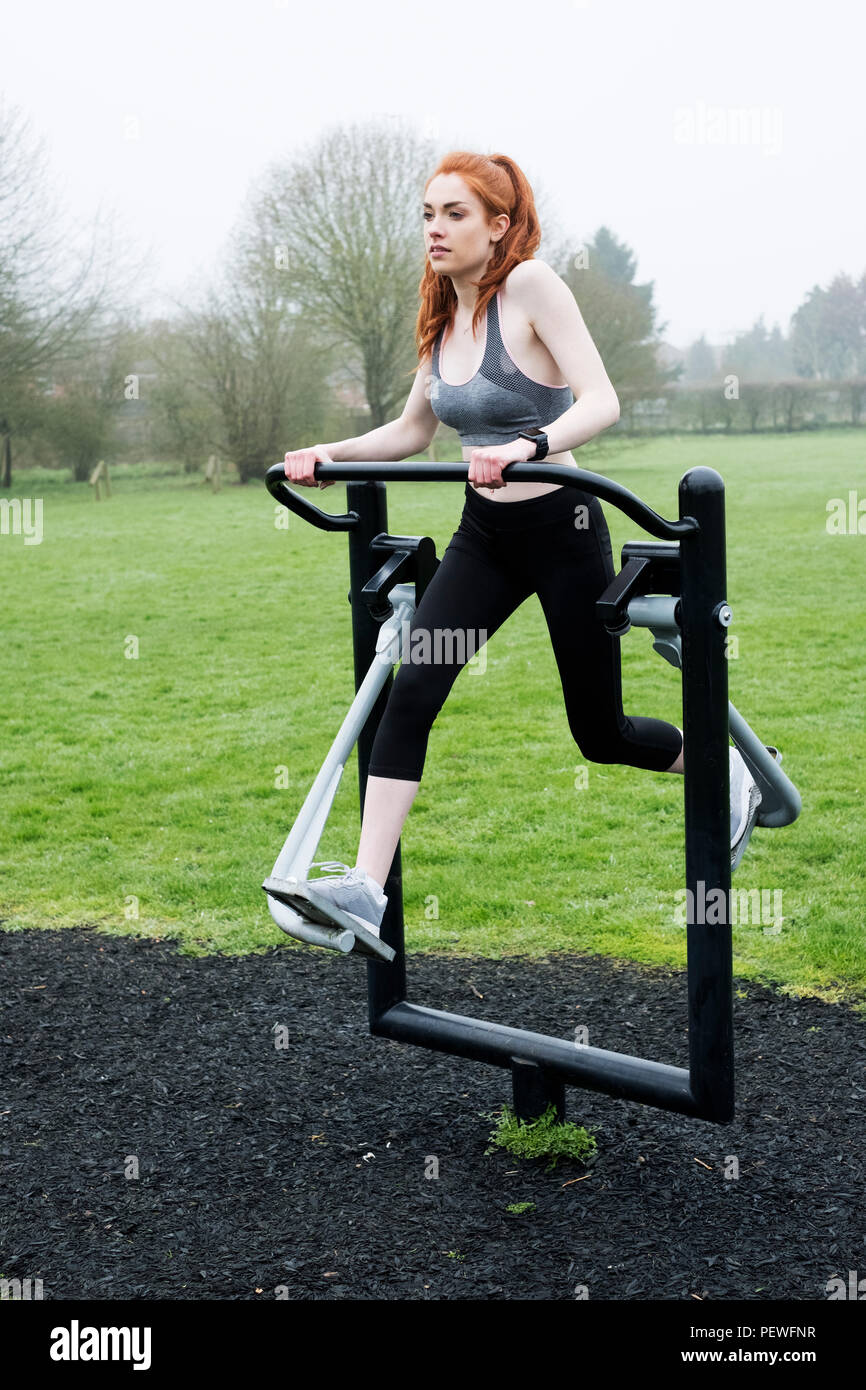 Young woman with long red hair wearing sportswear, using outdoor exercise machine. Stock Photo