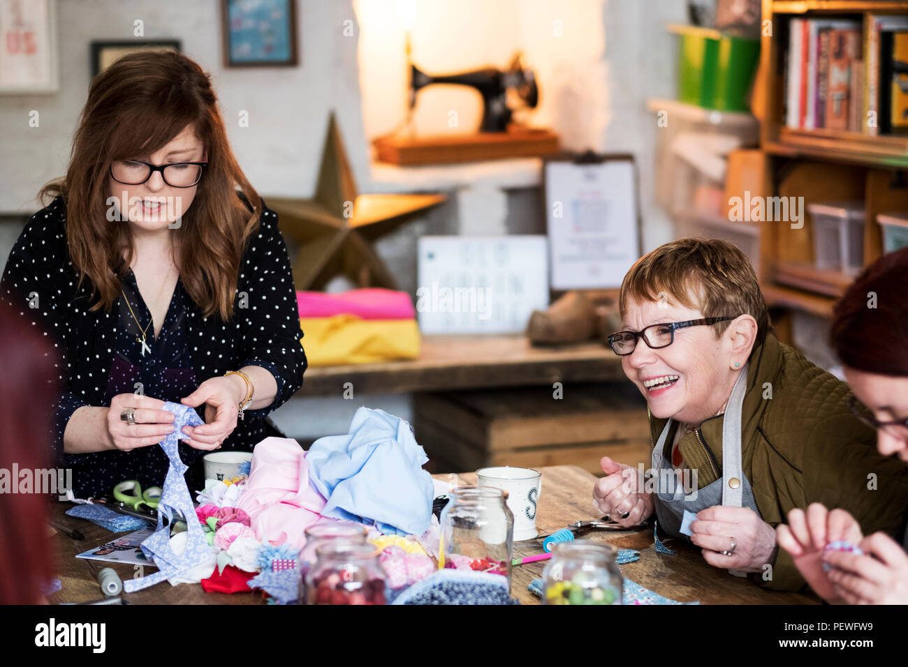 Two women sitting at a table in a workshop, making fabric flowers. Stock Photo