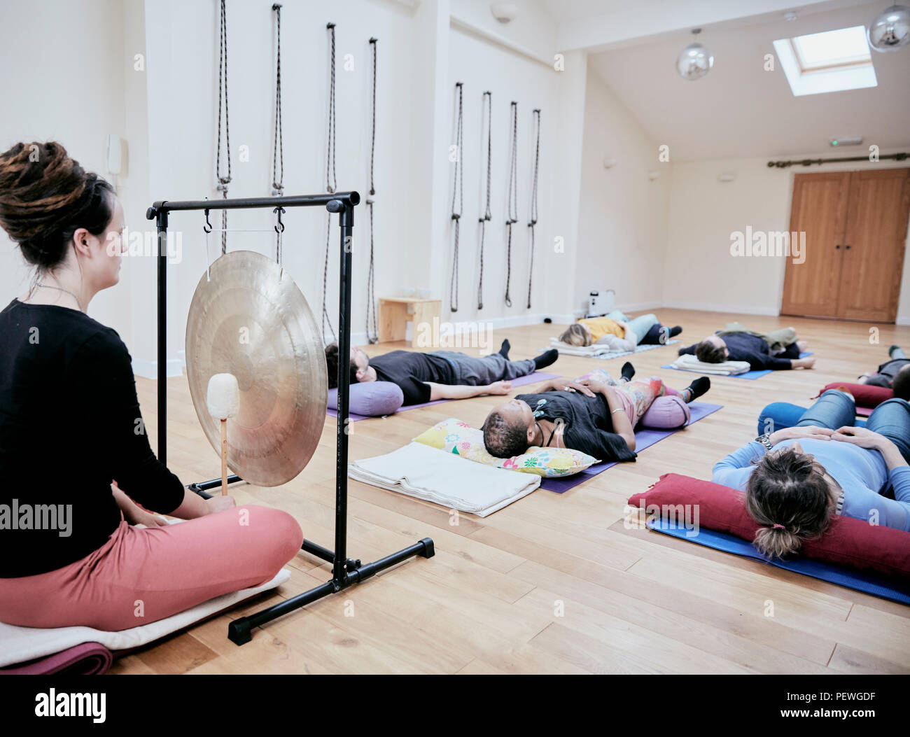 Woman using a gong during a sound therapy session and three people lying on their backs relaxing. Stock Photo