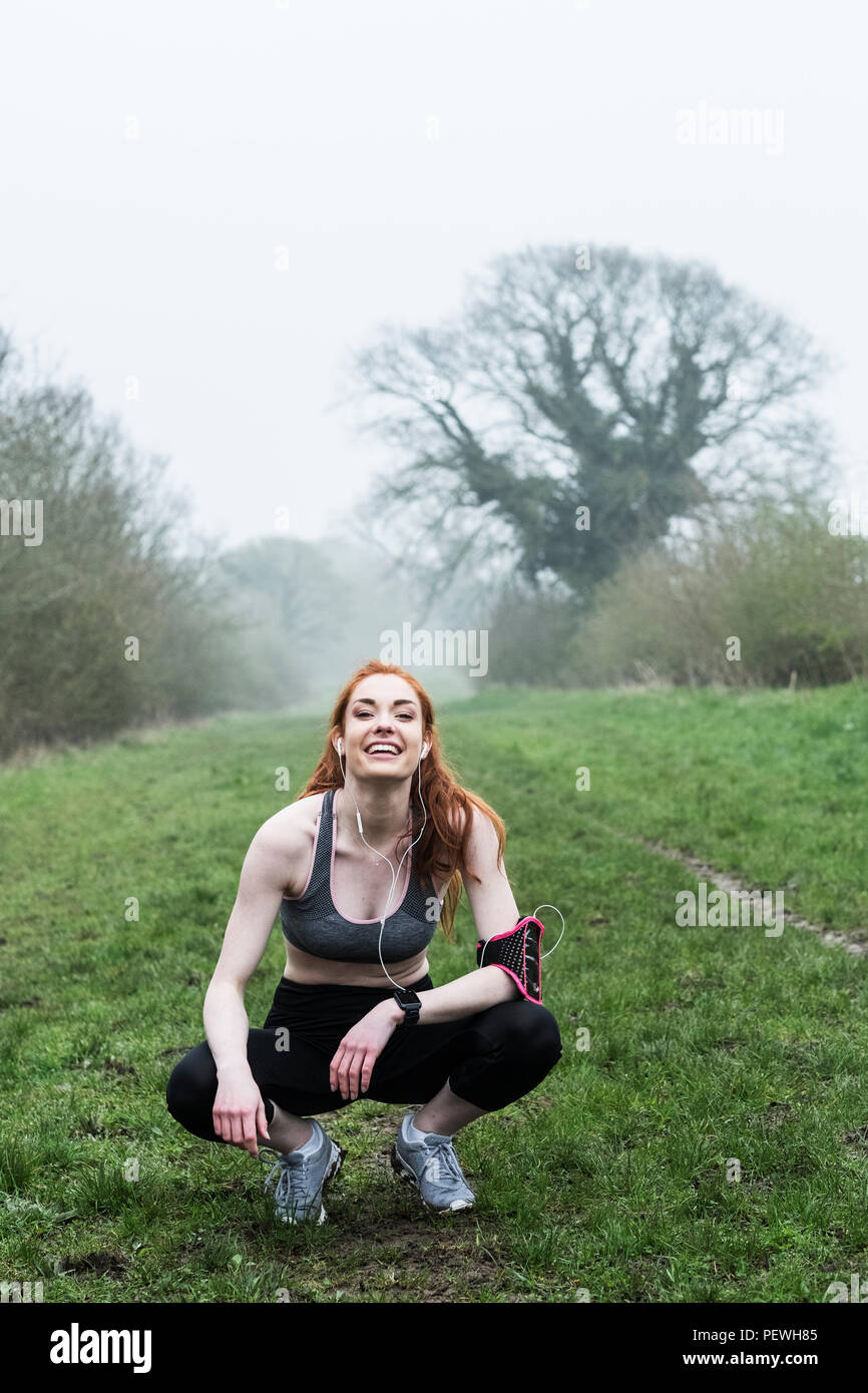 Young woman with long red hair wearing sportswear, resting, crouching down, outdoors, looking at camera. Stock Photo