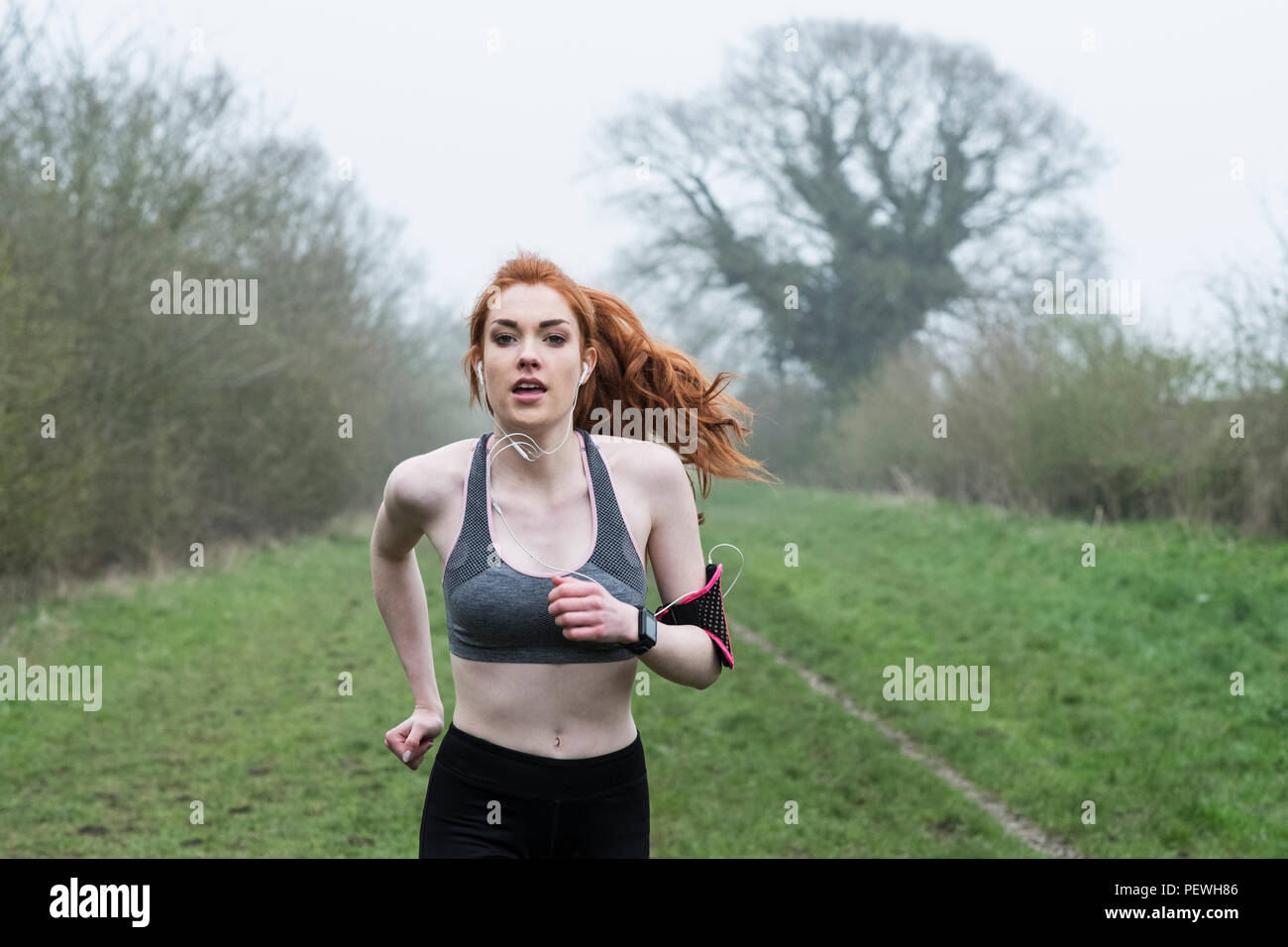 Young woman with long red hair wearing sportswear and earphones, exercising outdoors, running along, armband with music player, looking at camera. Stock Photo