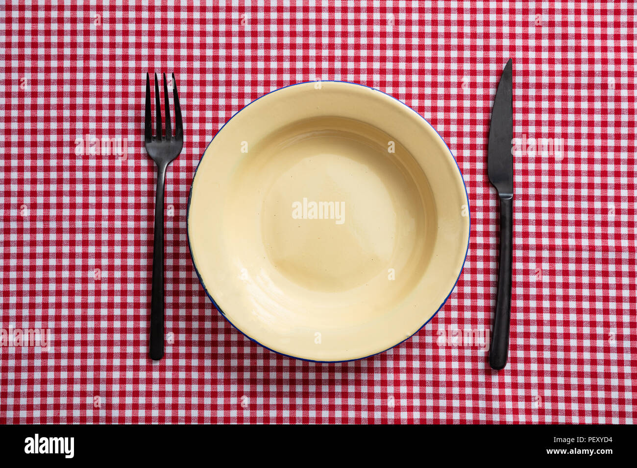 Rustic place setting. Empty enamel plate, fork and knife on red checkered tablecloth, top view Stock Photo