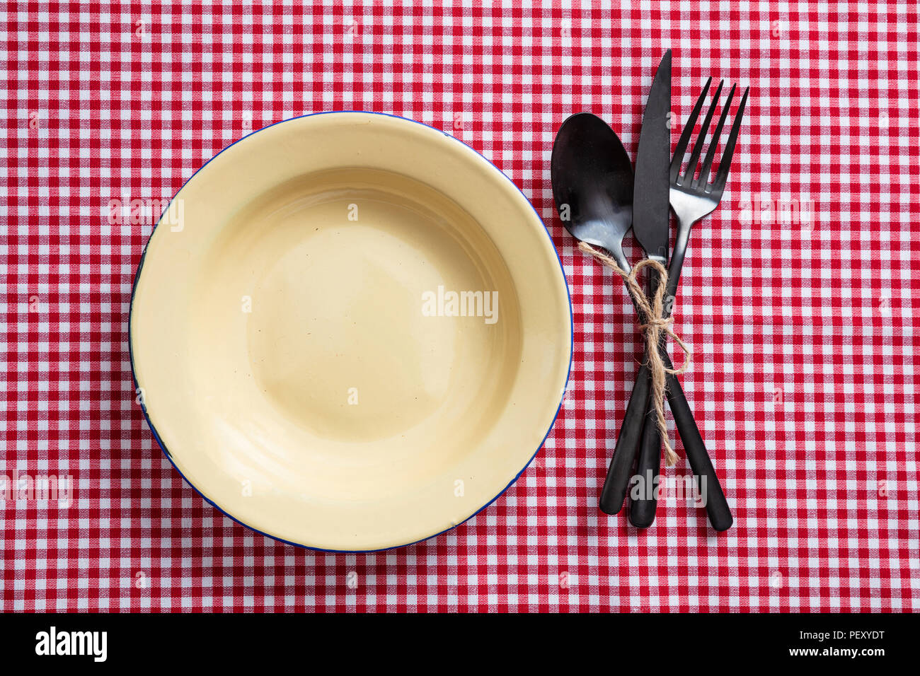 Rustic place setting. Empty enamel plate and cutlery on red checkered tablecloth, top view Stock Photo