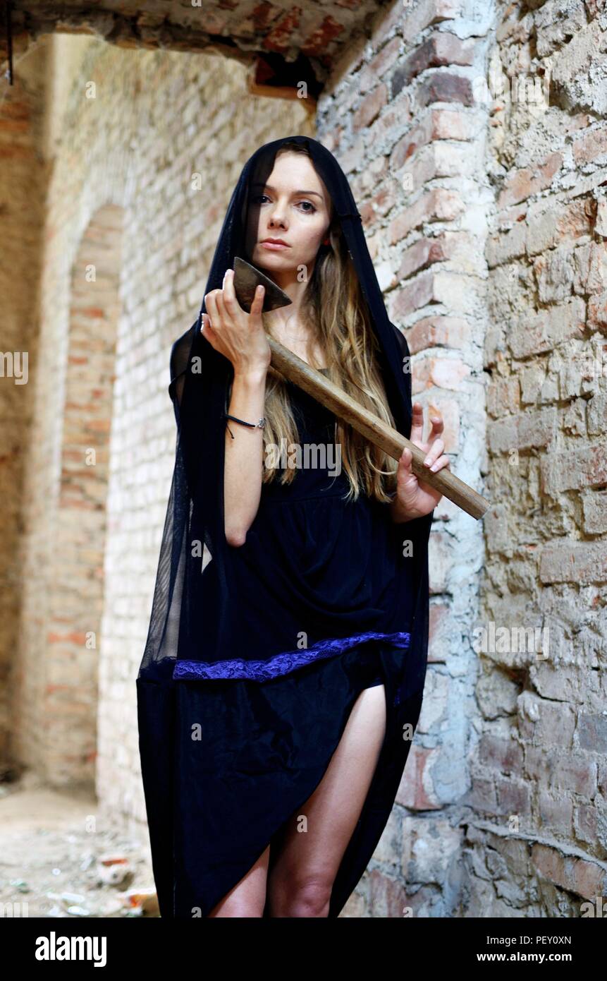Mysterious woman in the ruins holding an ax in her hand. Female model with black dress and covered head inside ruins, old palace in Poland. Stock Photo