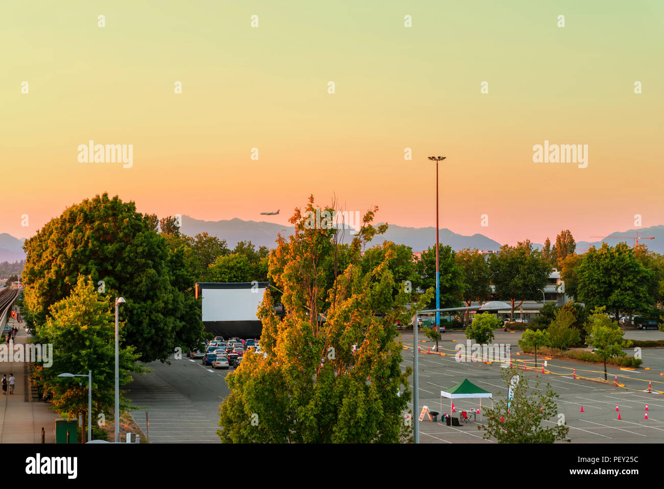 Car parking with summer cinema, green trees, mountains, flying plane, cars on summer evening in modern city Stock Photo
