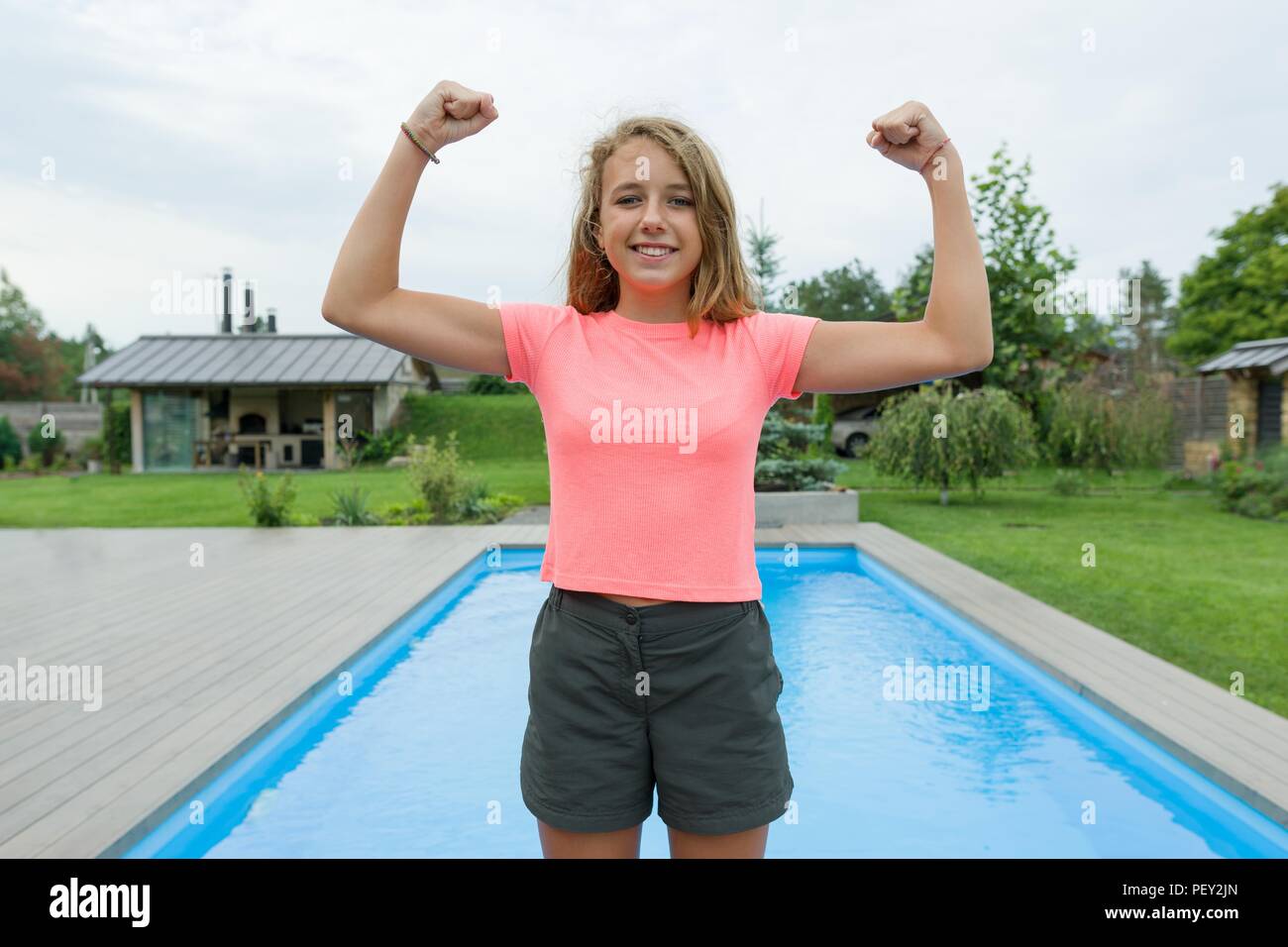 People, power, stamina, strength, health, sport, fitness concept. Outdoor portrait smiling teenage girl showing muscles, background swimming pool gree Stock Photo