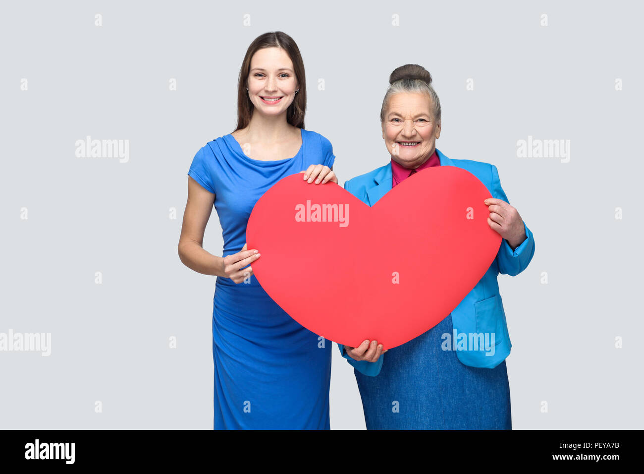 happy granddaughter and grandmother standing, holding big heart shape, looking at camera with toothy smile. Big love in family. Relations in the famil Stock Photo