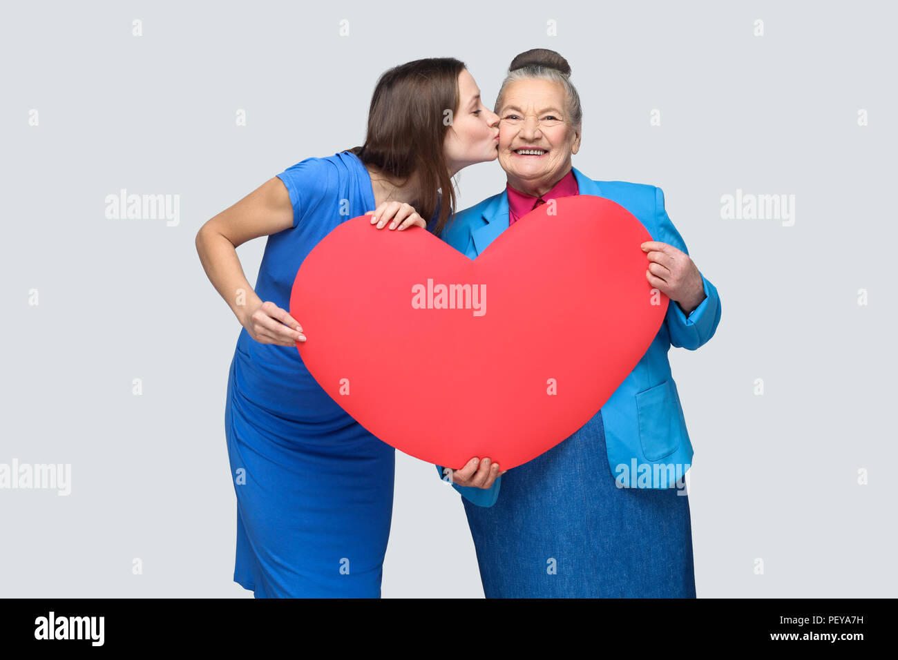 Granddaughter kissing grandmother standing, holding big heart shape, looking at camera with toothy smile. Big love in family. Relations in the family. Stock Photo