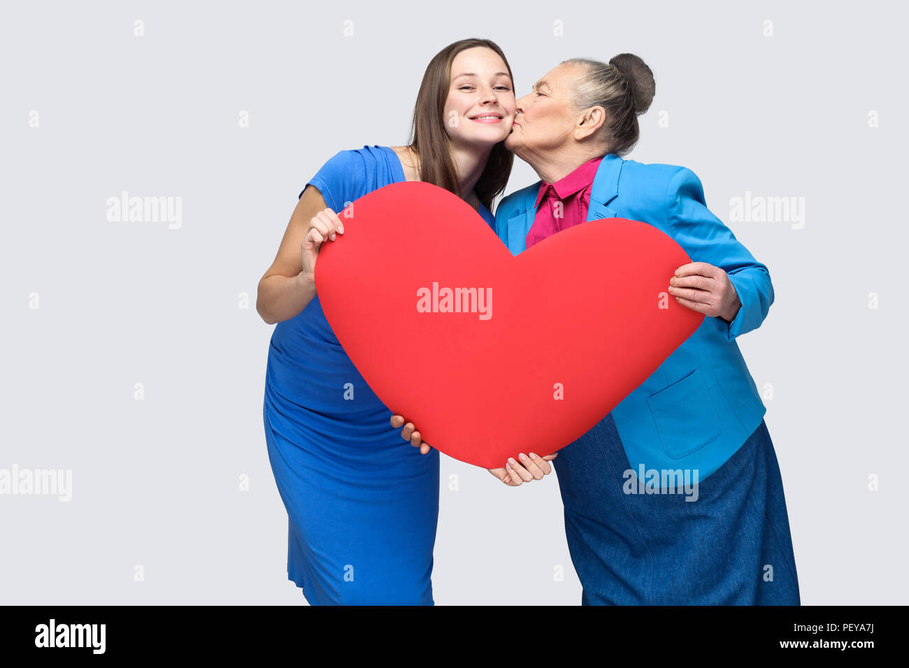 Grandmother kissing granddaughter standing, holding big heart shape, looking at camera with toothy smile. Big love in family. Relations in the family. Stock Photo