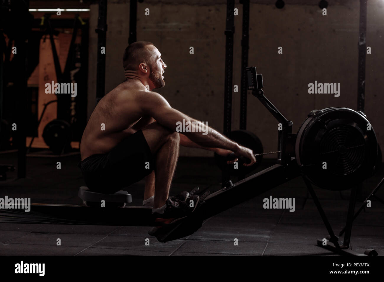 young handsome man is doing exercises with rowing machine indoors. side view full length photo Stock Photo