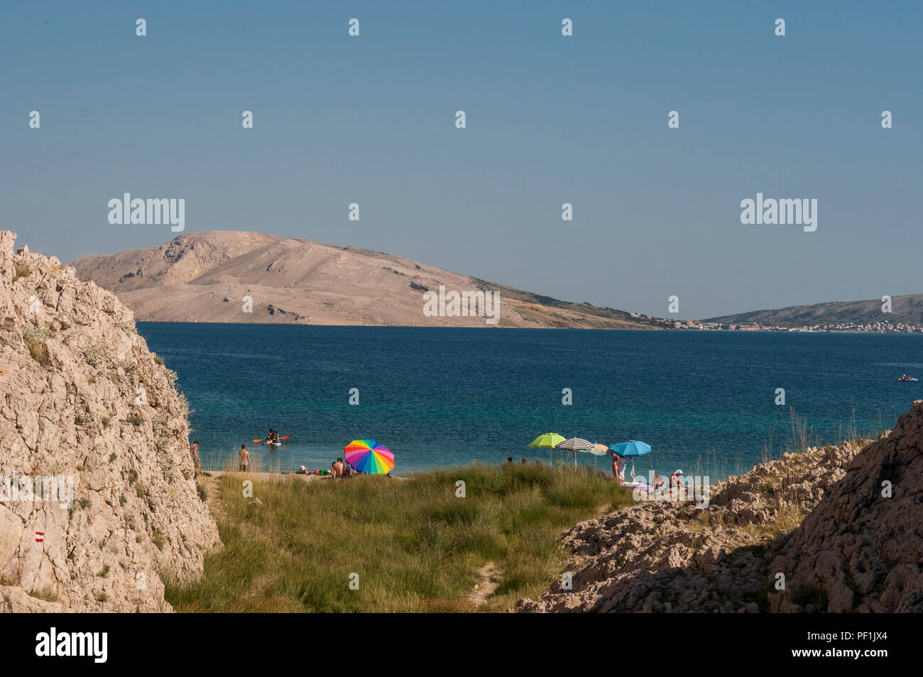 Pag Island, Croatia: rainbow beach umbrella in the grass at Rucica, pebbled beach nestled in a barren bay with green plants and desert landscape Stock Photo