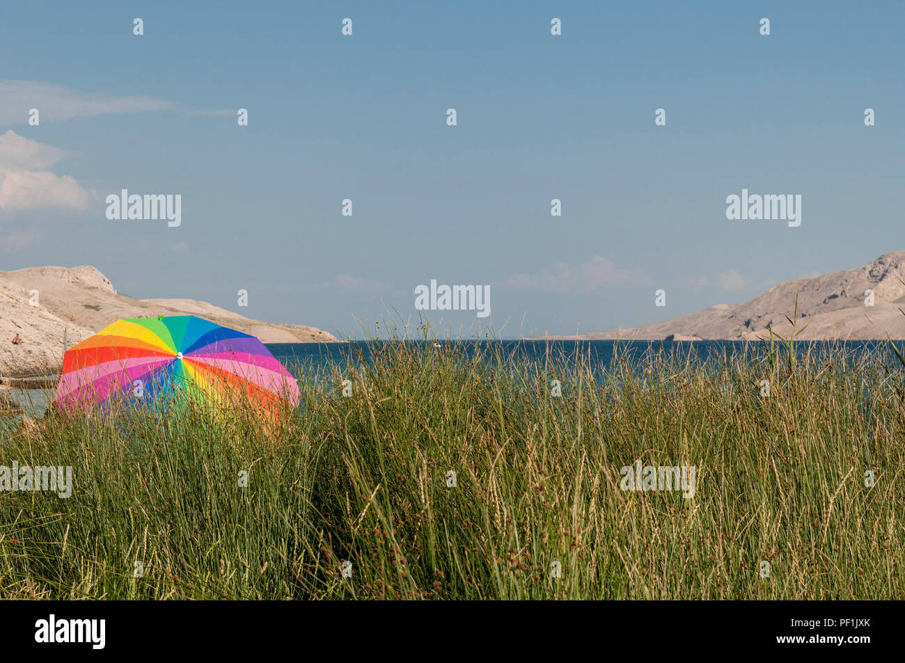 Pag Island, Croatia: rainbow beach umbrella in the grass at Rucica, pebbled beach nestled in a barren bay with green plants and desert landscape Stock Photo
