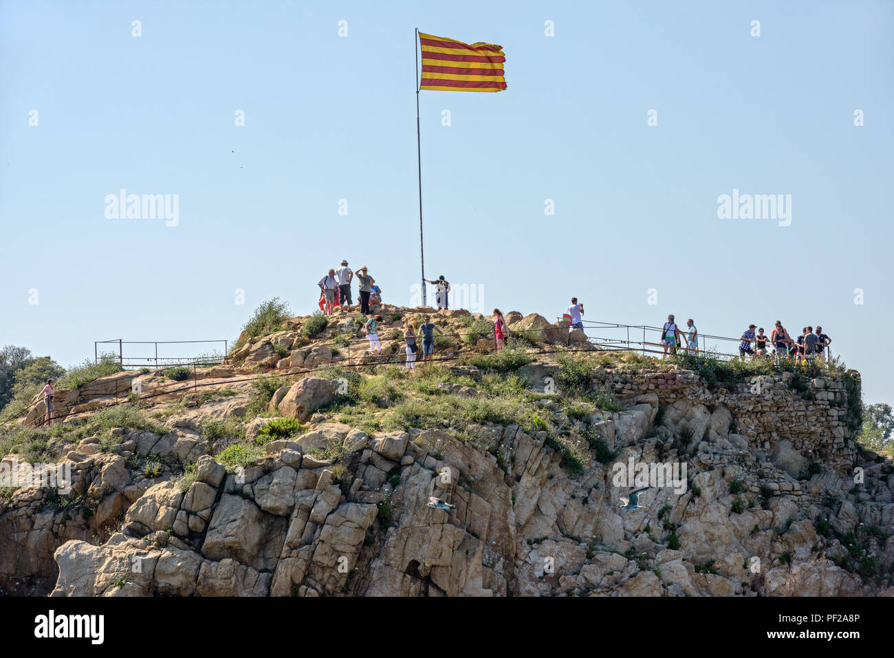 Tourists are near Senyera flag on the mast on top of Sa Palomera Rock in Blanes, Costa Brava coast in Catalonia, Spain. Stock Photo