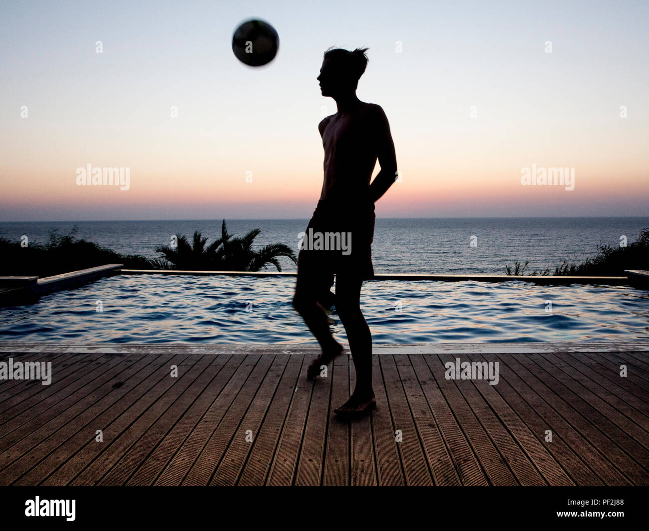 Model released picture of a boy practicing football tricks by a pool at sunset whilst his brother swims Stock Photo