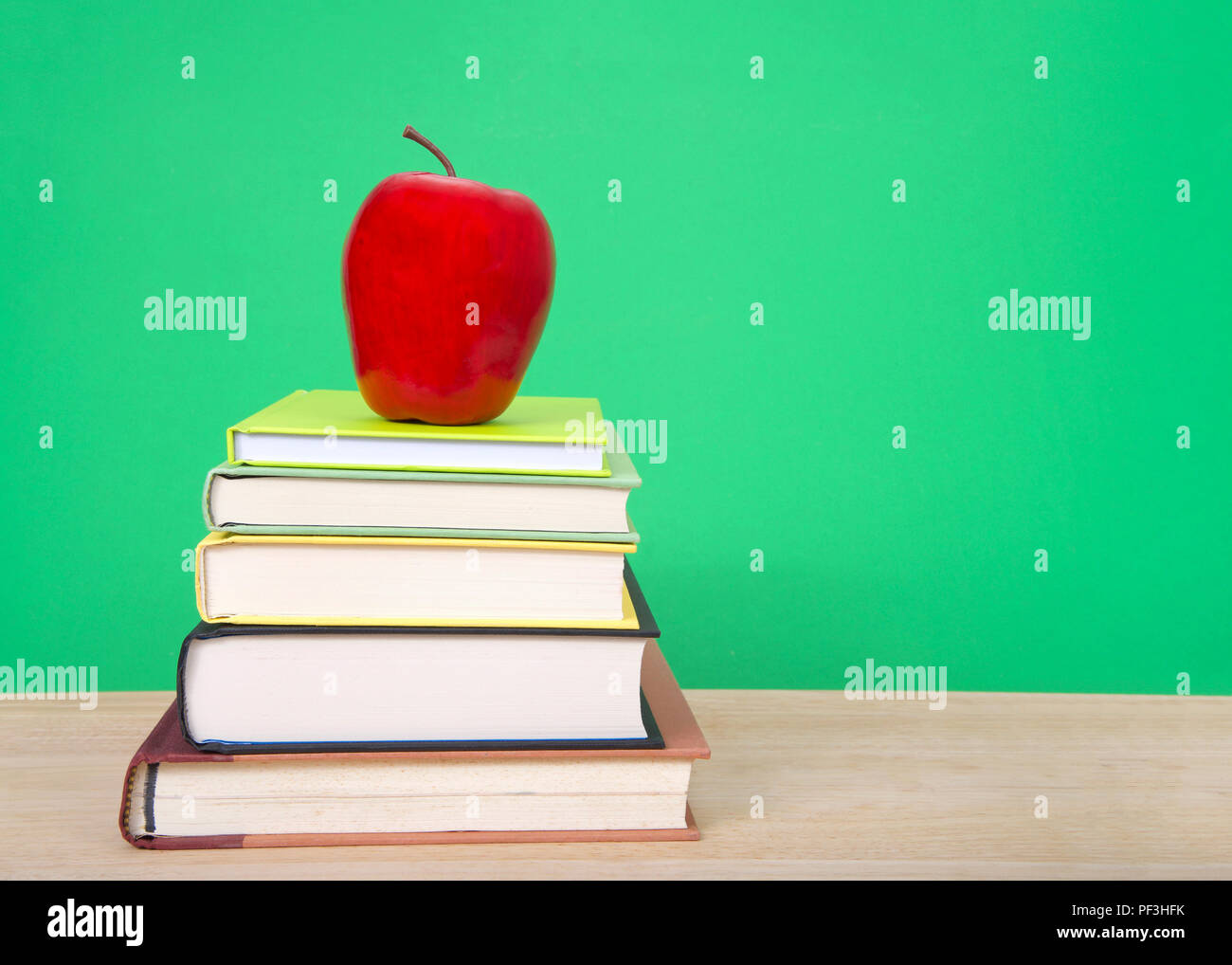 Hard bound books with various colored covers stacked on light wood table with red apple on top, dark green background similar to old fashioned chalk b Stock Photo