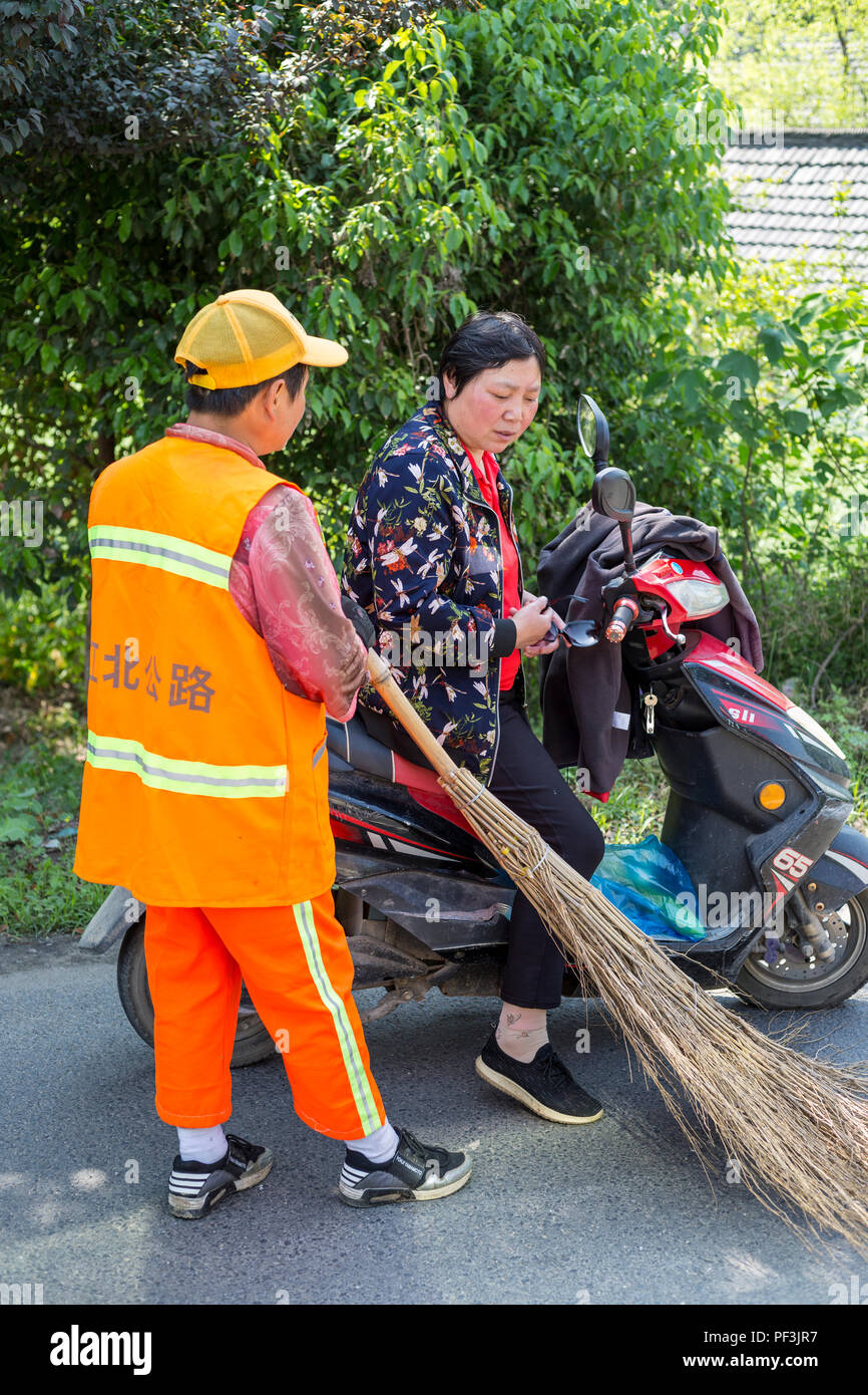 Yangzhou, Jiangsu, China.  Modernity Versus Tradition.  Street Sweeper with Traditional Broom, Woman Riding Electric Motorbike. Stock Photo