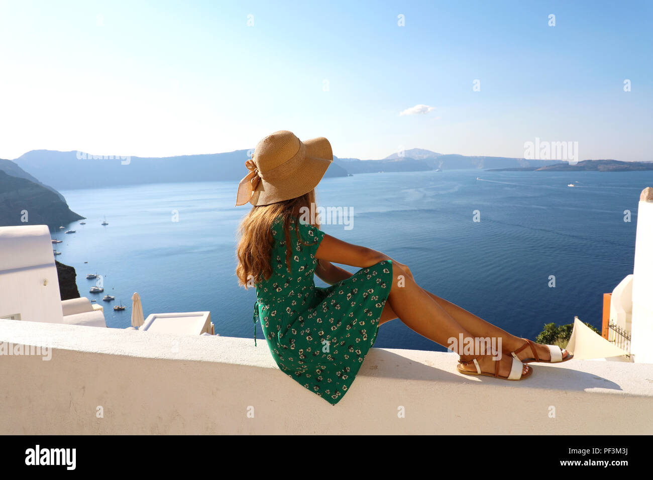 Beautiful young woman sitting on wall looking at stunning view of Mediterranean sea and Caldera in Santorini, Greece, Europe. Lifestyle woman Stock Photo