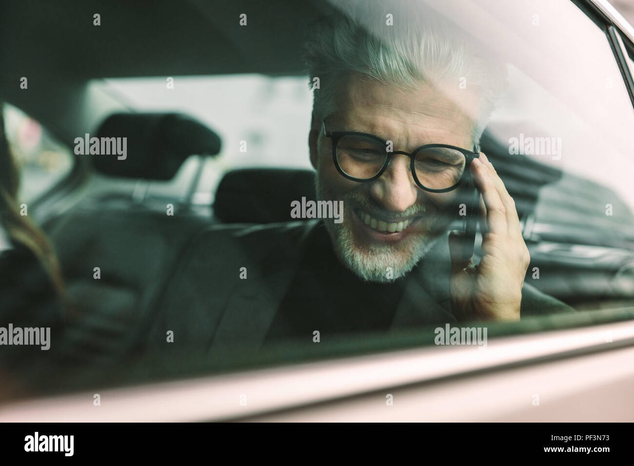 Smiling senior businessman making phone call while sitting on back seat of a car. Senior male commuter travelling by a cab and talking on phone. Stock Photo