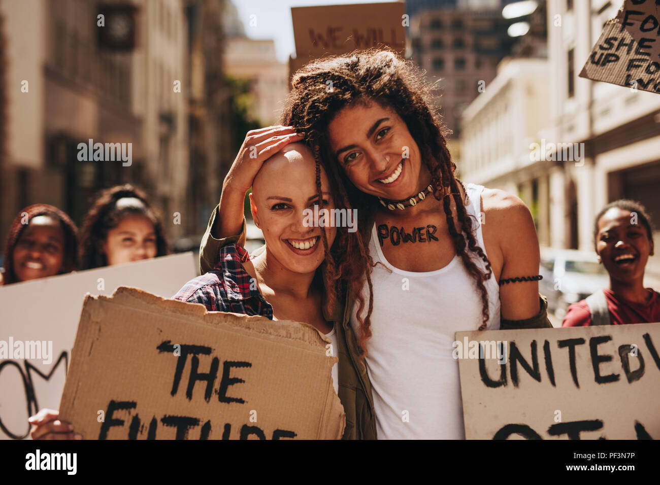 Portrait of two happy young woman with placards protesting outdoors with group of people in background. Stock Photo