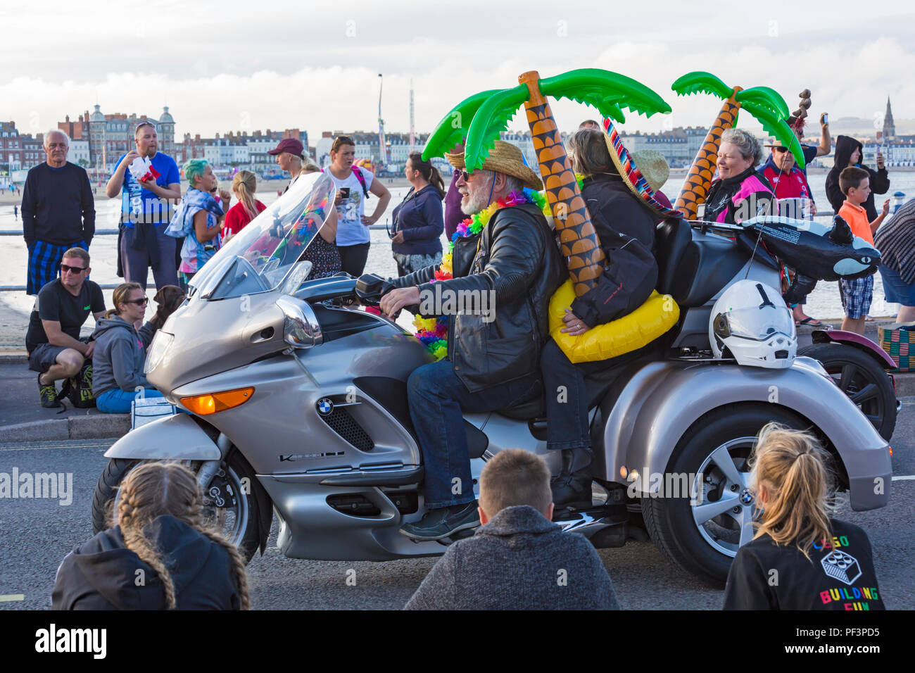 Couple riding with inflatable palm trees on BMW motorbike taking part in the annual carnival procession parade at Weymouth, Dorset UK in August Stock Photo