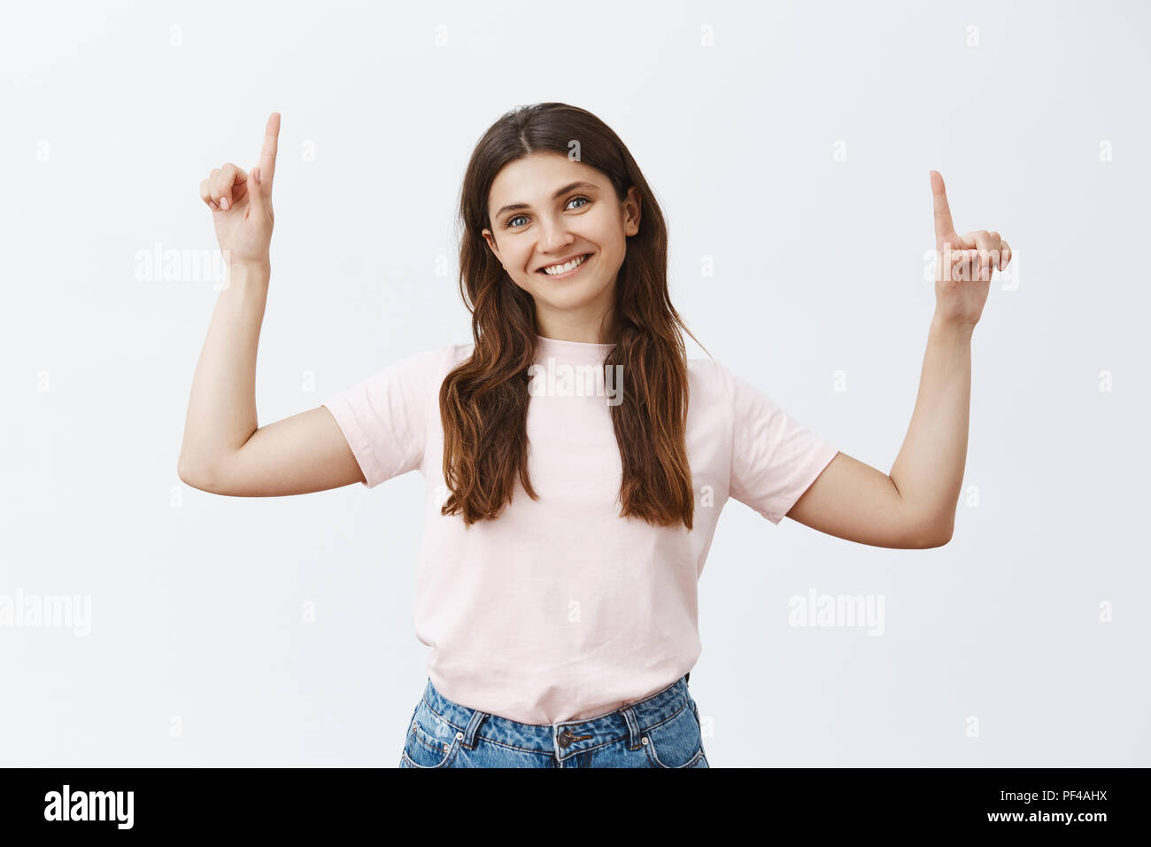 Woman graduated from university getting up corporate ladder. Portrait of pleased charming friendly-looking european girl in pink t-shirt raising hands pointing upwards and smiling with tilted head Stock Photo