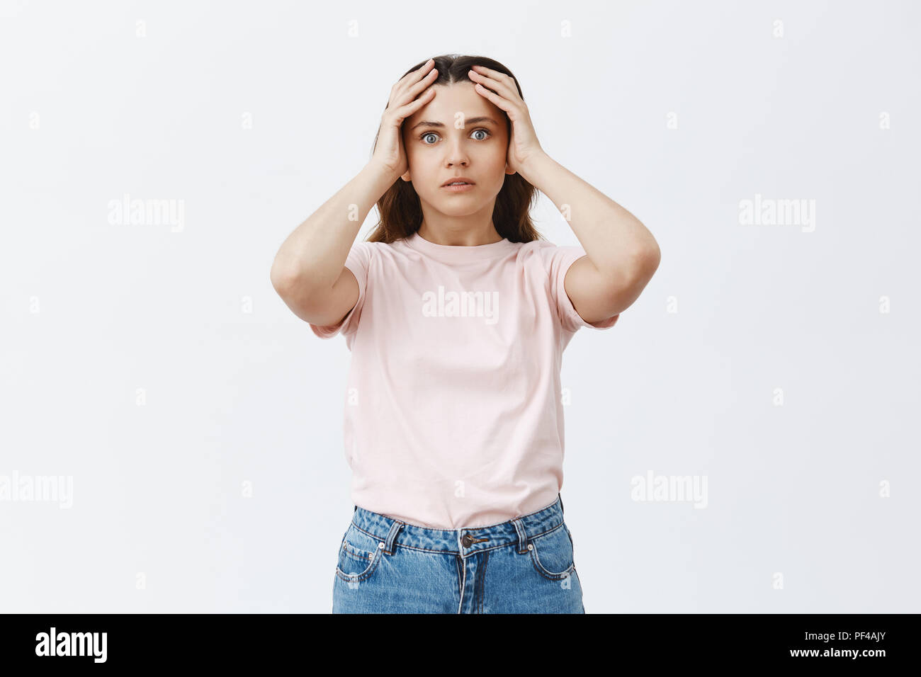 Woman facing troublesome situation being worried and tired. Portrait of stunned attractive young female studen in pink t-shirt holding hands on head and staring worried at camera Stock Photo