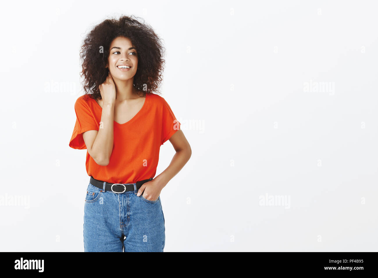 Indoor shot of carefree good-looking dark-skinned woman with afro hairstyle touching neck and looking right with charming smile, feeling little nervous before important meeting over grey wall Stock Photo