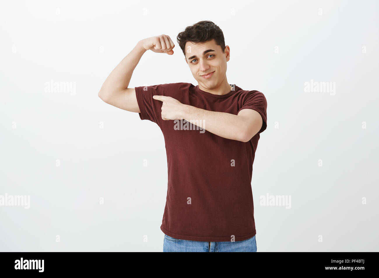 Portrait of handsome masculine happy guy in red t-shirt, raising hand and showing muscles, pointing at arm with index finger, bragging about body strength after visiting gym, enjoying working out Stock Photo